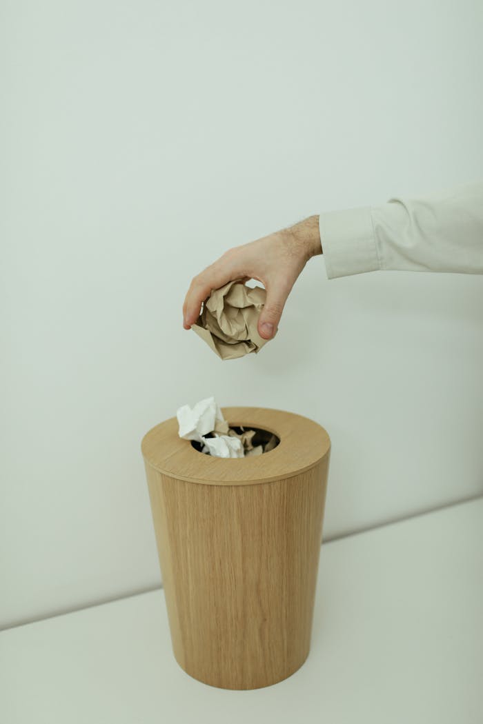 A hand throws crumpled paper into a simple wooden trash bin indoors.
